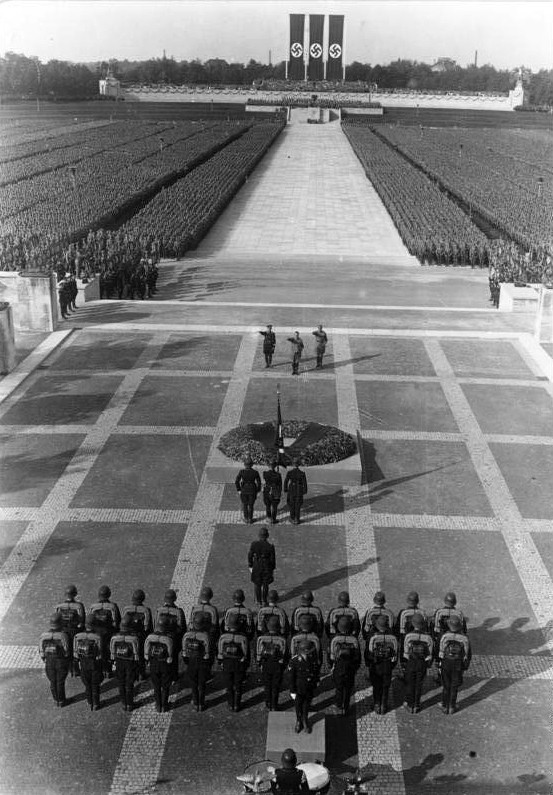 Totenehrung (hommage aux morts) lors du rassemblement de Nuremberg en 1934. Le chef SS Heinrich Himmler, Adolf Hitler et le chef SA Viktor Lutze (de gauche à droite) sur la terrasse en pierre devant la Ehrenhalle (salle d'honneur) de la Luitpoldarena. À l'arrière-plan, on peut voir la Ehrentribüne en forme de croissant. | Archives fédérales allemandes